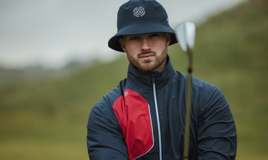 A golfer is using his club to measure his next shot. He is wearing a rain bucket hat with a Galvin Green logo on, and a navy golf rain jacket with red details. 