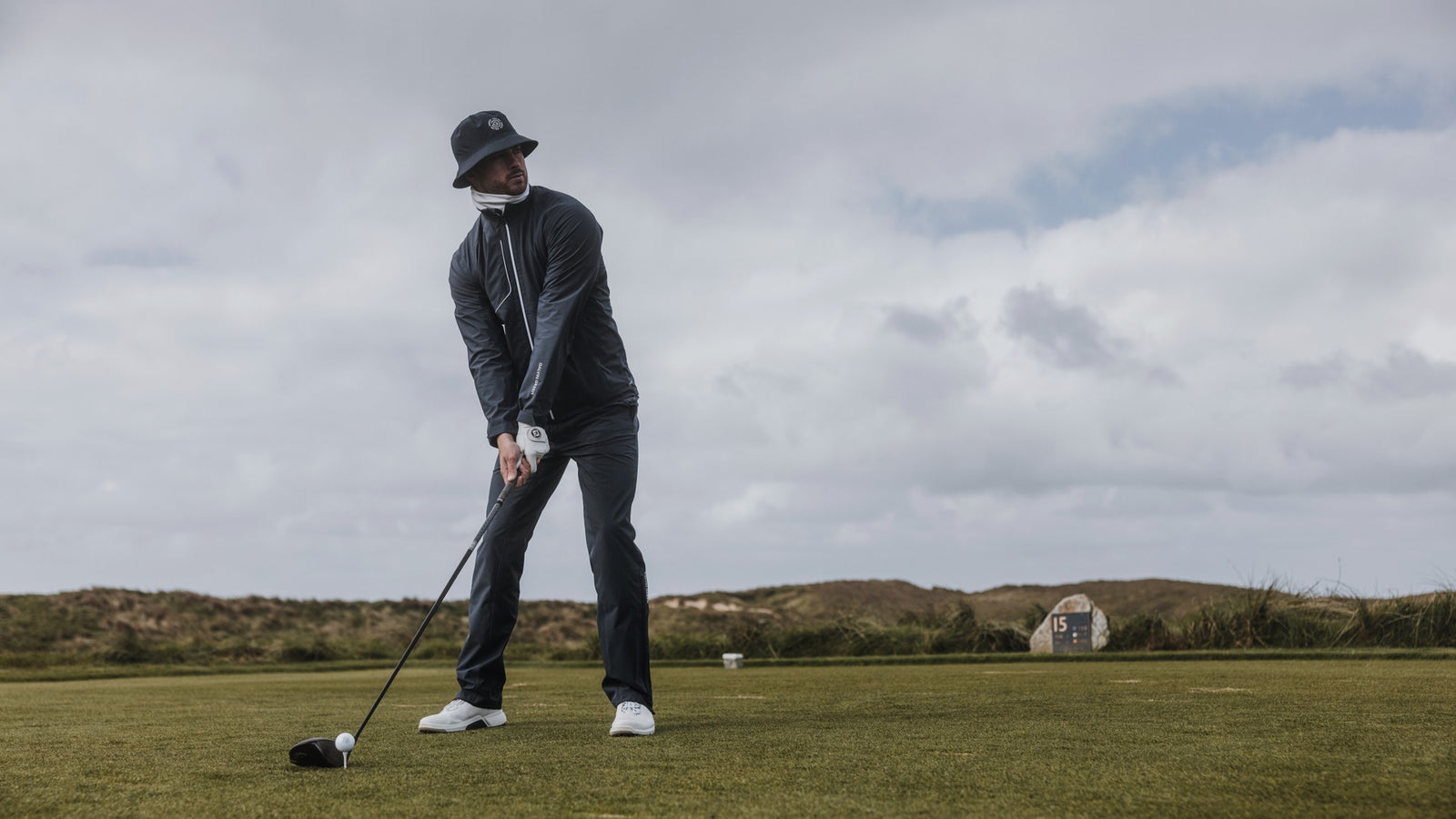 A focused golfer in a navy waterproof golf jacket, pants and waterproof golf hat is about to tee off.