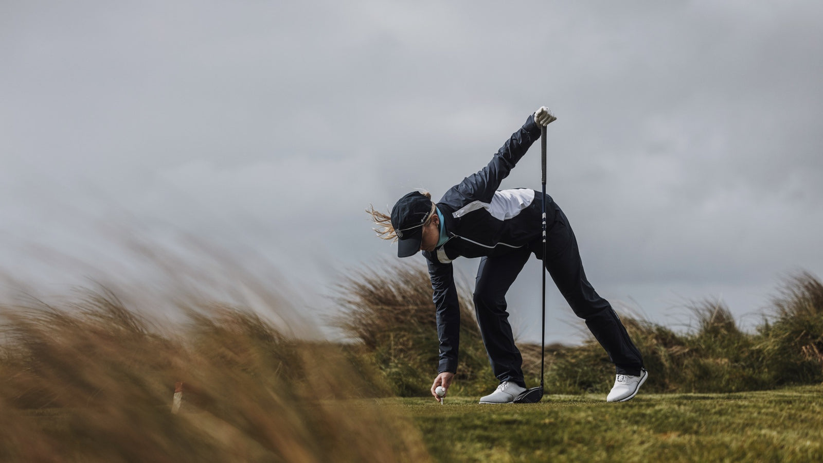 A female golfer is placing a golf ball on a golf tee. It's windy outside. She is waring a waterproof and windproof golf jacket, cap and golf pants in navy.