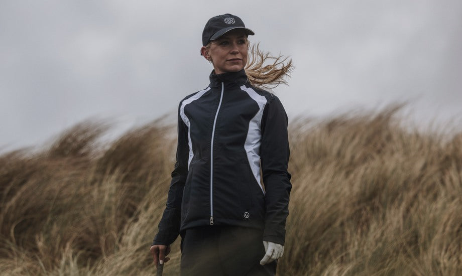 A woman is standing on very windy golf course. You can see the wind in her hair and in the tall grass behind her. She is wearing a waterproof golf hat and a black rain golf jacket with white details. 