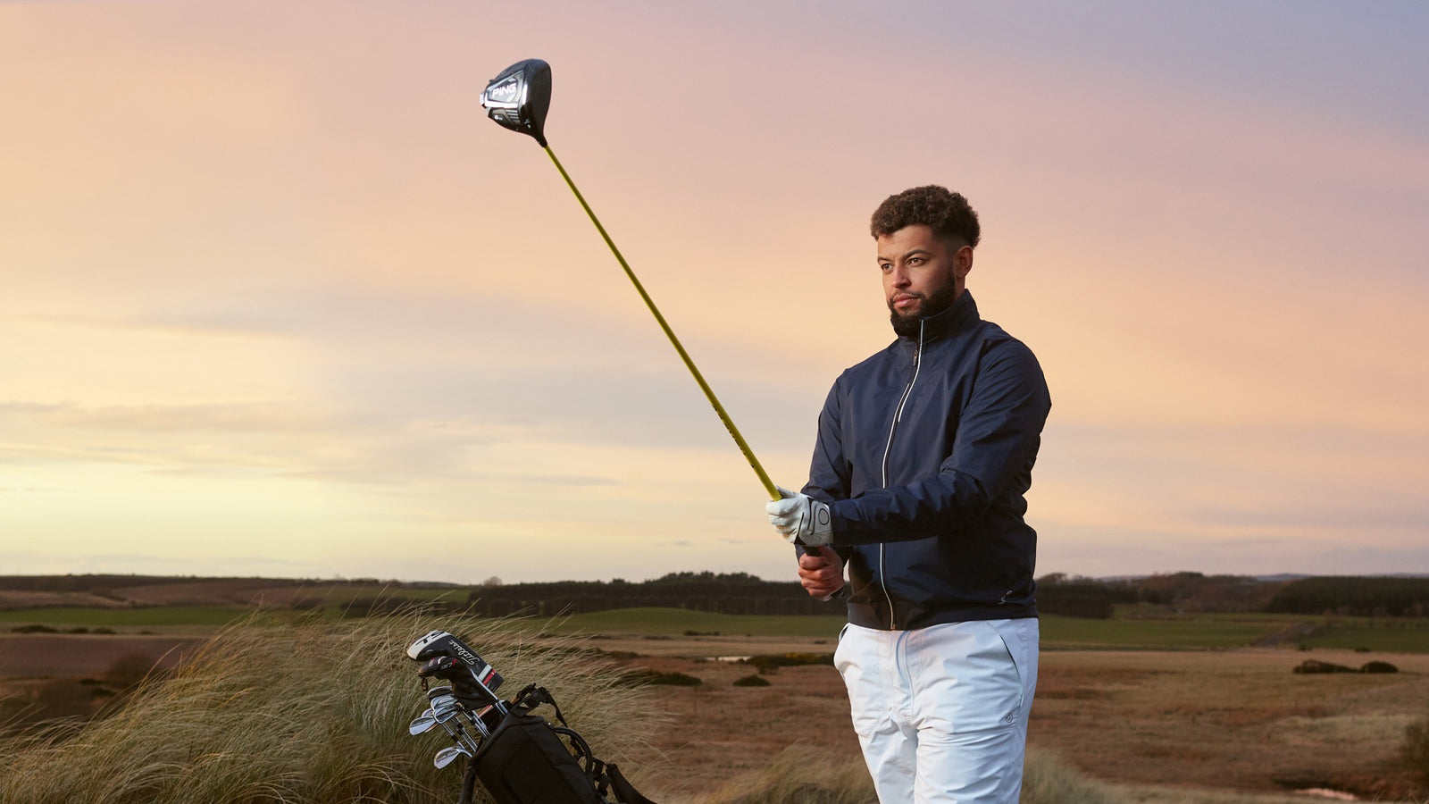 A golfer is aiming with his driver in the sunset. He is wearing a golf rain jacket and waterproof pants. 