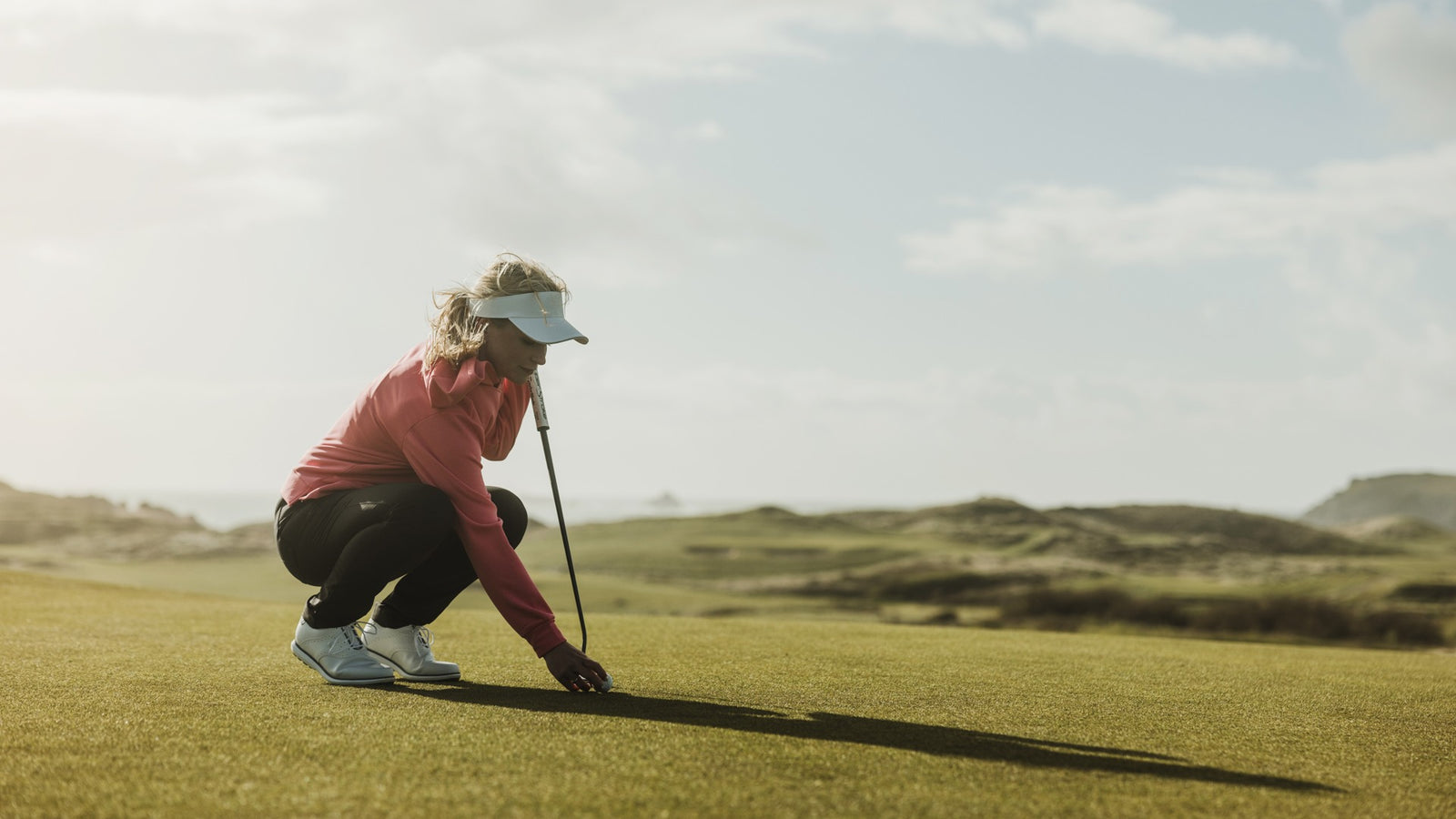 A woman is placing a golf ball on the green and preparing for her next shot. She is wearing a sun visor, a pink golf mid layer and stretchy golf pants.