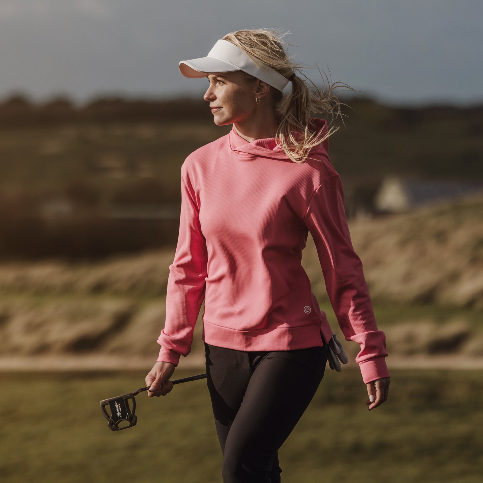 A female golfer is walking on a sunny golf course. She is wearing a pink mid layer and a white visor.