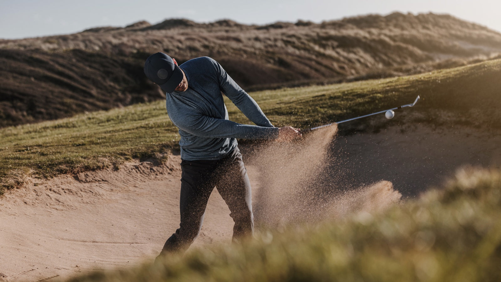 a golfer is hitting the ball in a bunker. He is wearing a blue golf mid layer and a matching navy cap.