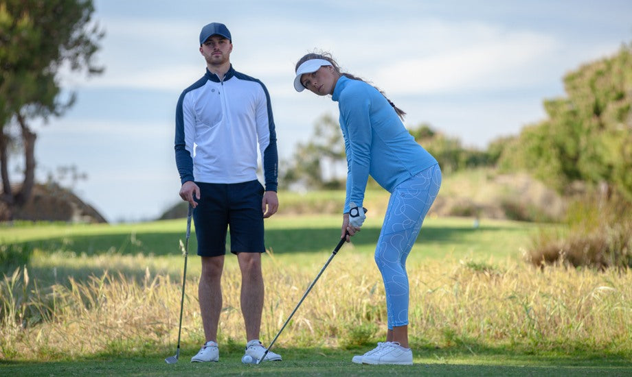 A man and a woman on a golf course. She is about to hit the ball and is wearing a golf mid layer and matching stretchy golf bottoms. He is standing behind her and is wearing a golf sweater in white and navy, a navy golf hat, and navy golf shorts. 