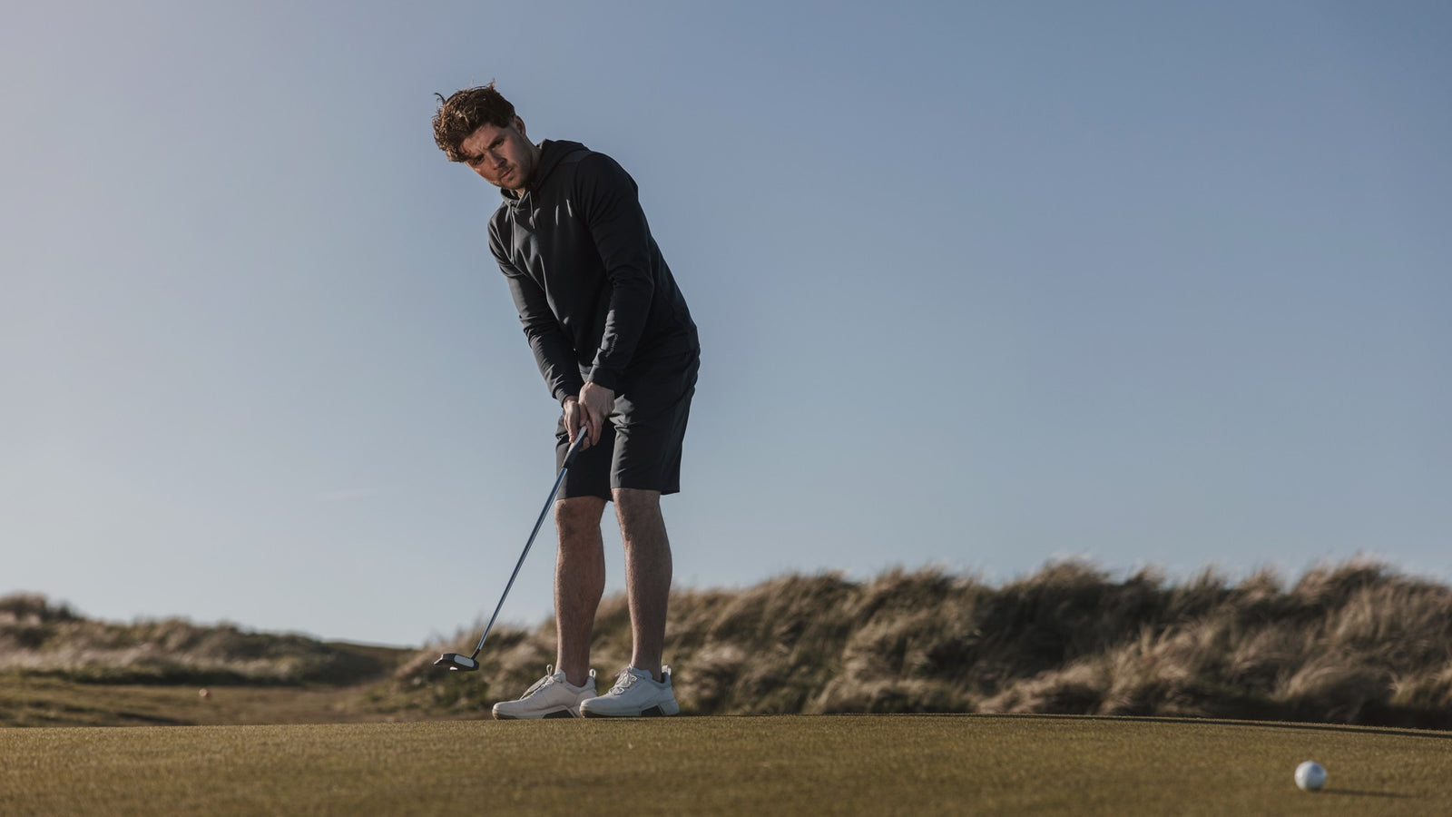 The golfer has just been putting on the green. He is wearing a black golf hoodie and golf shorts. 