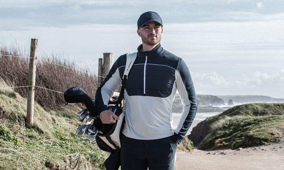 A man, who is wearing a padded half zip sweater in white and navy, is standing by the sea carrying his golf clubs. 