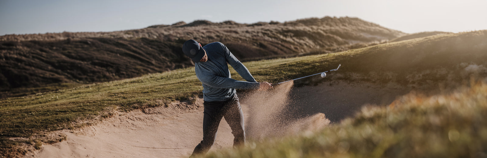 A golfer is hitting a ball out of a bunker. He is wearing a golf hat with a round Galvin Green logo.