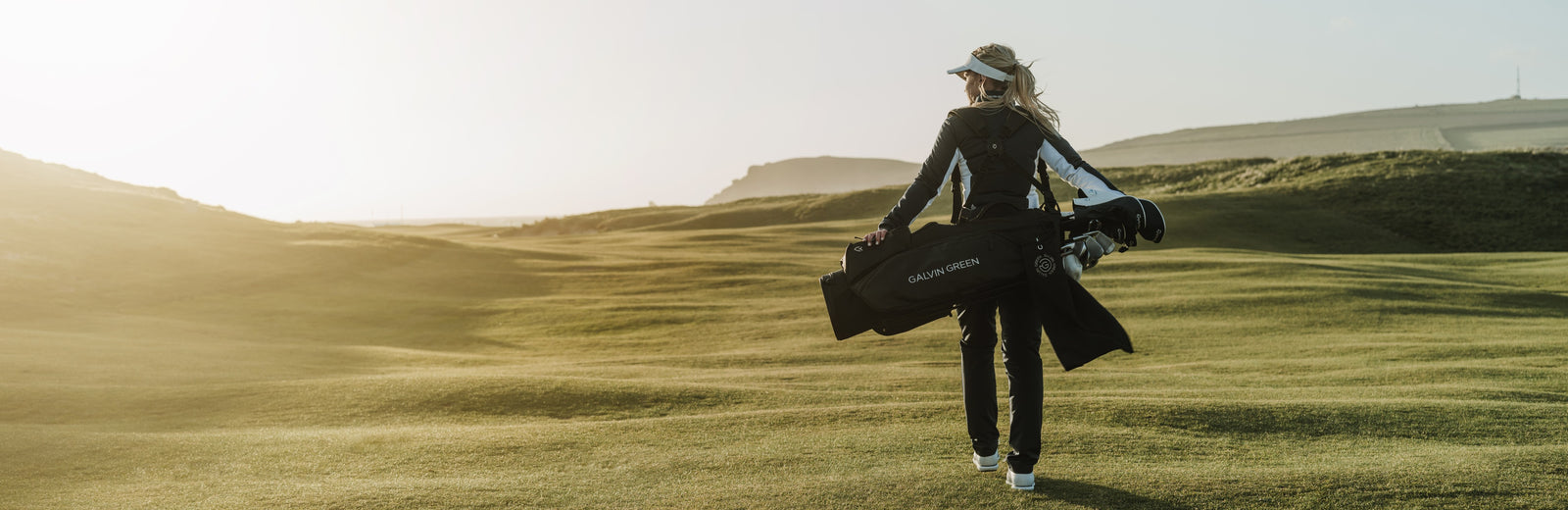 A woman is walking on an empty golf course with her bag over the shoulder. Because how the light falls, it looks like it's early in the morning. She is wearing a black and white golf jacket, black golf pants, and a white golf shade. 