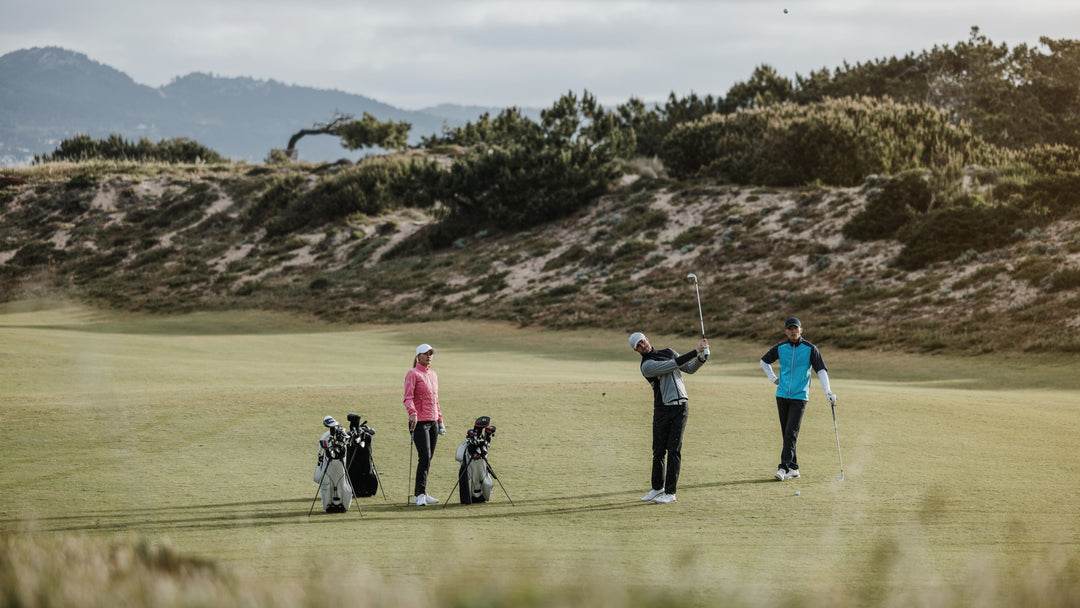 Three golfers on a golf course. The woman is wearing a pink padded windbreaker, the man hitting the ball is wearing a black and grey wind breaker, and the man watching is wearing a short sleeve golf jacket.