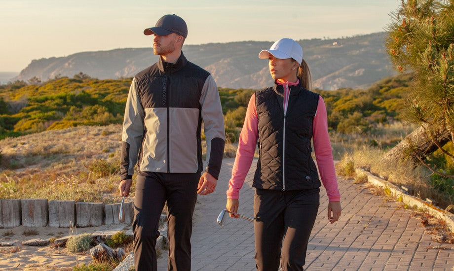 A man and a woman are walking on a walkway on a nice golf course. He is waring a windproof golf jacket, a grey golf cap and black pants. She is wearing a pink mid-layer, a padded golf vest, and a white golf hat. It looks like this sun is about to set.