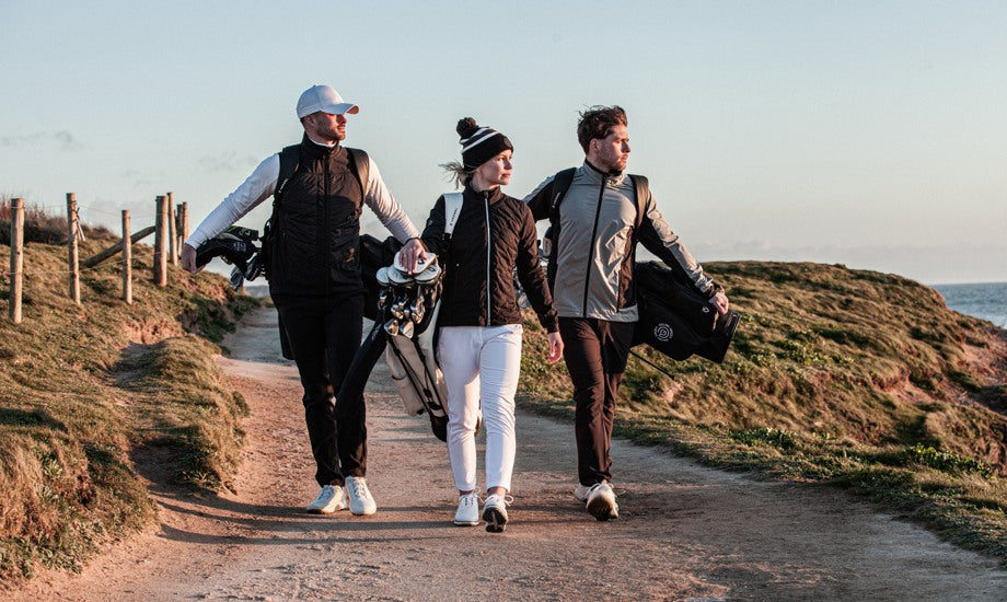 Three golfers are walking on a windy path carrying their clubs. They are wearing wind proof jackets, golf pants, and golf hats.