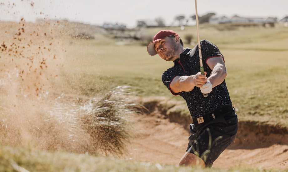 A golfer is hitting the ball in a bunker, the sand is splashing everywhere. He is wearing a printed golf polo shirt, shorts and a red golf hat. 