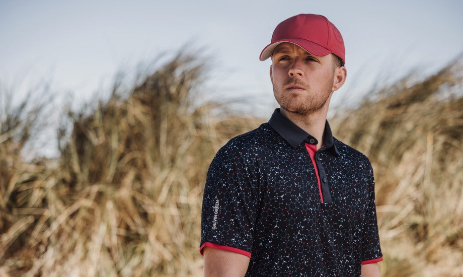 A man who is wearing a red golf cap and a printed breathable golf polo with red details is looking into the distance. There is tall grass behind him. 