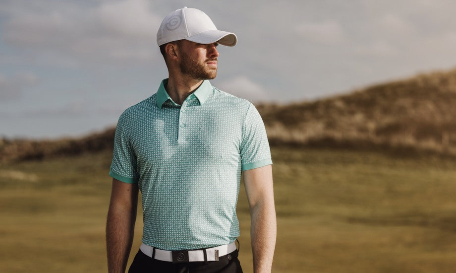 A bearded man in a green golf polo shirt is standing on a golf course. He is wearing a breathable hat and is looking into the distance. 