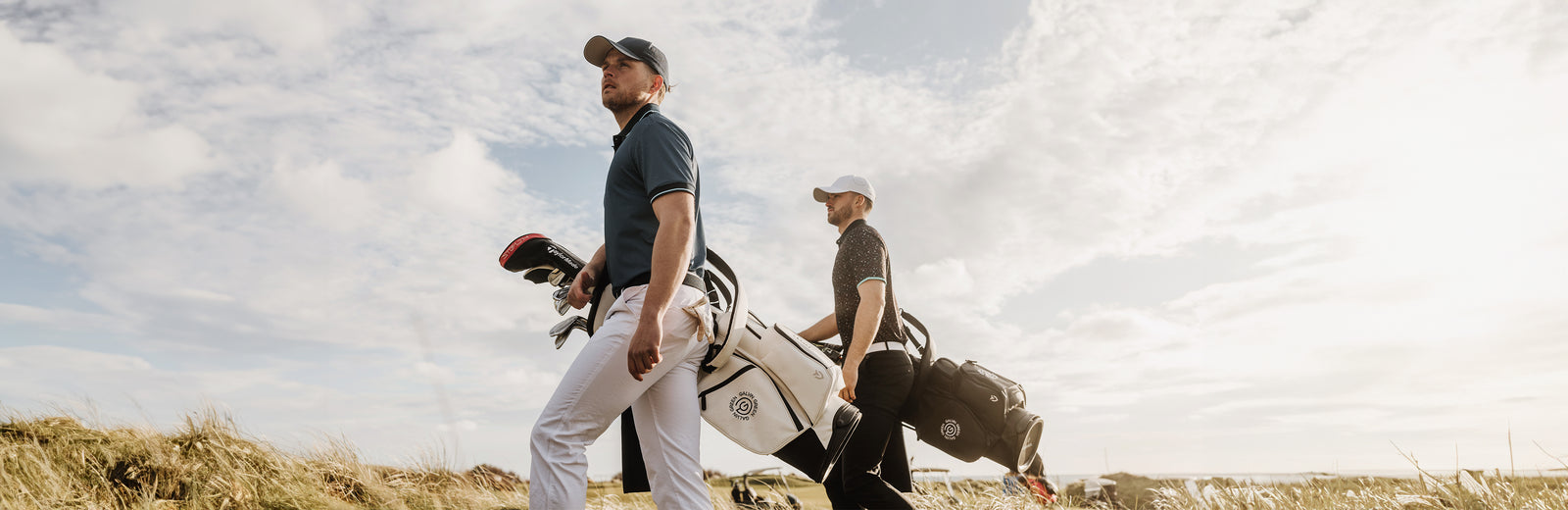 Two men in golf shirts, hats and golf bags are walking on a sunny golf course. 